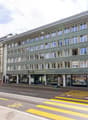 Multi-story building, green facade, glass windows, shops on the ground floor, bicycle parking sign
