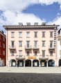This image shows a row of multi-story buildings with arched entrances and balconies in a European-style town square. The buildings have a mix of orange, pink, and white facades, and there is a Coop supermarket visible on the ground floor of one of the bui