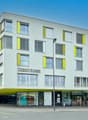 A modern, multi-story commercial building with a gray exterior and yellow accents. The building has large windows and a prominent Credit Suisse logo displayed on the facade. There are retail spaces on the ground floor and offices or apartments on the uppe