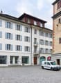 This image shows a historic town square with a tall clock tower and several multi-story buildings with traditional Swiss architectural styles. The buildings have white facades with blue shutters and balconies. There is a cobblestone plaza in the foregroun