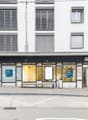 The image shows the exterior of a UBS bank branch. The building has a white facade with horizontal striped windows. The entrance is located in the center, with the UBS logo displayed prominently. There are several glass doors and windows, allowing visibil