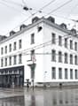 A multi-story white building with ornate architectural details, including windows and a prominent roofline. The building appears to be located in an urban area, with power lines and other infrastructure visible overhead. There are bicycles parked outside