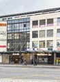 Multi-story commercial building with various retail stores and businesses, including Migros, Axa, Akros, and Denner. The building has a modern glass and concrete facade with signage for the different companies. There are parking spaces in front of the bui