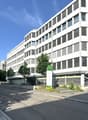 Multi-story office building with a modern, angular design featuring large windows and louvered facades. The building has a prominent green sign labeled 'Block 22' on the ground floor. There are trees and landscaping around the building, and a paved road i