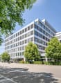 Raised ground floor, office building, several floors, many windows, hedges and trees in front, street with parked car