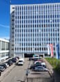 multistory building, numerous glass windows, open parking lot, Swiss flags, white and blue exterior