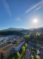 The image shows an aerial view of a town or city surrounded by mountains and forests. The town has a mix of residential and commercial buildings, with a river or stream running through the center. There are cars parked on the streets and trees lining the