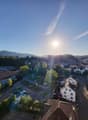The image shows a panoramic view of a small town or city surrounded by hills and mountains. The town has a mix of residential and commercial buildings, with trees and greenery visible throughout. The sky is clear and blue, with the sun shining brightly. T
