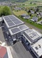 Large building with solar panels on the roof, multiple parking spaces, surrounded by houses and greenery