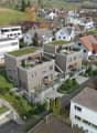 Multi-story apartment building with balconies, raised ground floor, and parking spaces. The building has a modern, minimalist design with a mix of white and gray colors. The surrounding area appears to be a residential neighborhood with other houses and b