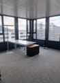 This image shows a modern office space with large windows providing a view of the surrounding cityscape. The room has a minimalist design with a white table, a black office chair, and a gray carpeted floor. The ceiling features a grid-like pattern of pane