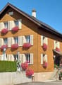2 story house, brown wooden exterior, white shutters, pink flowers in window boxes, garden with hedges and plants, awning over entrance