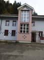 Two-story building, white and pink facade, multiple windows, red entrance door, brown roof, located on a road