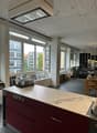 An empty office space with large windows, large sink, kitchen island, wooden table and chairs, kitchen cabinets, and racks with various items. There is a large glass window, with a view of the buildings outside. The room is mostly empty, with several furn
