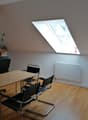 Attic office space with wooden beams, large skylight window, wooden desk, black office chairs, and hardwood flooring.