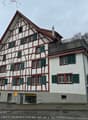 White painted, timber-framed house, red roof, multiple windows, red trims, small garden