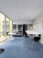 Large open office space with floor-to-ceiling windows, gray carpeted floors, white walls, and modern furniture including a sofa, tables, and chairs. The space appears to be designed for a professional work environment.