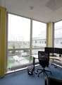 Spacious office with large windows providing a view of the outdoor scenery, including trees and buildings. The office is furnished with a desk, computer, and office chairs. The walls are painted in a light color scheme, and the floor is covered with blue