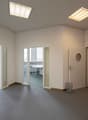 Empty room with white walls, gray carpeted floor, glass doors and windows, round window in door, trash can, desk with chair in the background, ceiling lights