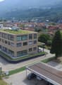 Aerial view of a building with a green roof, surrounded by a parking lot with several cars parked, a road with cars driving, and a mountain in the background.