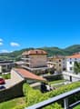 View from a balcony with a railing, overlooking a street with buildings, mountains, a car, and a tree.
