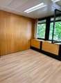 The image shows a spacious office room with wooden paneled walls, a wooden floor, and large windows overlooking a green outdoor area. The room has a clean and modern design, with built-in storage units along the walls.