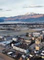 Aerial view of a city surrounded by mountains, with a lake or river in the foreground. The city appears to have a mix of residential and industrial/commercial buildings.