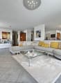 Spacious open-concept living room with large sectional sofa, glass coffee table, and artwork on the walls. The room has a modern and bright design with tile flooring and recessed lighting.