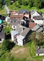 Aerial view of a small village nestled in a hilly, forested area. The village consists of traditional stone and tile-roofed buildings, some raised off the ground, with narrow streets and paths winding through the landscape.