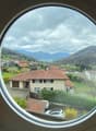 The image shows a scenic view through a round window. It depicts a mountainous landscape with a small town nestled in the valley below. The town has residential buildings with red tile roofs, surrounded by lush greenery and trees. In the distance, there a