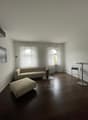 large sofa, foot stool, round table, floor lamp, two large arched windows with curtains, picture frame, hardwood floor, ceiling lamp, white walls
