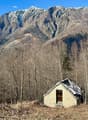 Small stone cabin in a forested mountain setting, with snow-capped peaks in the background