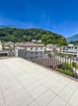 Spacious balcony with tiled flooring, metal railing, overlooking a residential area with buildings, trees, and mountains in the background.