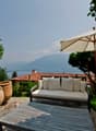 The image shows an outdoor patio area with a comfortable sofa, a large umbrella, and two large terracotta pots. The patio overlooks a scenic landscape with mountains in the background, suggesting a beautiful view. The patio is surrounded by lush greenery,
