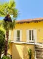 Two-story house, yellow exterior, red tile roof, balcony on the first floor, windows with white shutters, steps leading up to the entrance, front yard with palm tree and greenery.