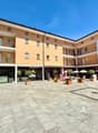 Modern multi-story building with a paved courtyard, balconies, multiple entrances, and trees.