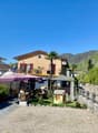 A building with two floors, beige exterior, outdoor dining tables, plants, flags, a small house, mountains, and a clear sky.