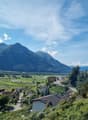 The image shows a scenic mountain landscape with a village nestled in the valley below. The mountains in the background are tall and rugged, with a blue sky dotted with clouds overhead. The village consists of a cluster of buildings with red-tiled roofs,