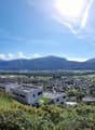 The image shows a panoramic view of a town or city surrounded by mountains. The town has a mix of residential and commercial buildings, with a modern-looking building visible in the foreground. The landscape is lush and green, with trees and vegetation co