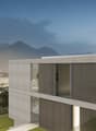 Modern concrete and glass building with balconies, situated on a hillside overlooking a city with mountains in the background. The building has a minimalist, geometric design with horizontal and vertical lines. There is a red tree in the foreground, addin