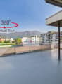 The image shows a 360-degree virtual tour of a balcony or terrace overlooking a city. The balcony has a concrete floor and a wooden roof structure. The view includes several multi-story buildings in the distance, with mountains visible in the background.