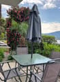 Outdoor patio with a glass-topped table and chairs, surrounded by lush tropical plants and flowers. There is a closed umbrella and a view of the mountains and lake in the background.