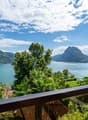 The image shows a scenic view of a lake surrounded by mountains and lush greenery. There is a balcony or terrace in the foreground, with palm trees and other tropical vegetation visible. The sky is blue with fluffy white clouds, creating a picturesque and