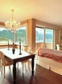 Spacious open-plan living and dining area with large windows offering a scenic view of the mountains and lake. The room features a wooden dining table with chairs, a plush sofa, and a decorative chandelier. The decor has a warm, cozy feel with neutral ton