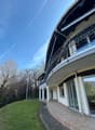 A curved, multi-story building with a balcony or terrace, surrounded by a grassy area and trees.