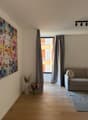 an empty living room with white walls, brown floor, two white vases, one with flowers, gray sofa, rug and large window