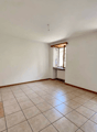 This image shows a spacious room with white walls and a tiled floor. The room has large windows that provide natural light and a view of the outdoors. The room appears to be empty, with no furniture or decor visible.