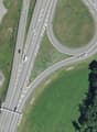 The image shows an aerial view of a highway interchange with multiple lanes and ramps. There is a grassy area in the center of the interchange, surrounded by trees and other vegetation. Several vehicles can be seen on the highway. There appears to be a bu