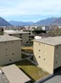 The image shows a residential area with several multi-story apartment buildings. The buildings have a beige exterior and are surrounded by paved areas and small grassy spaces. There appears to be a parking area visible in the foreground. The buildings are