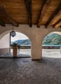 open area with wooden beams ceiling, cobblestone floor, wooden bench, potted plant, two arched openings leading to outside view